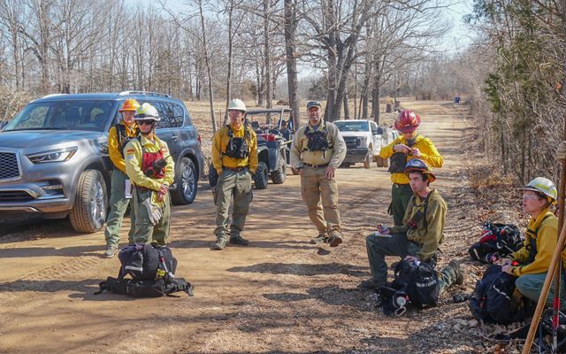 A small group of people in fire gear stand on a rural forested dirt road.