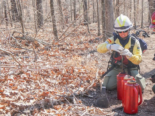 Kylie Paul and Lindsey Reinarz in fire gear kneel in a forested landscape working on two red drip torches.