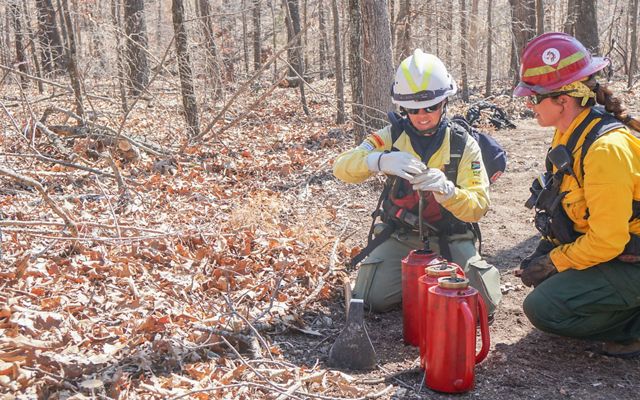 Kylie Paul and Lindsey Reinarz in fire gear kneel in a forested landscape working on two red drip torches. 