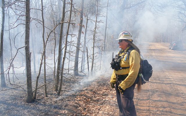 Kelly Martin stands in full fire gear along a dirt road with smoke from a prescribed fire in the background..