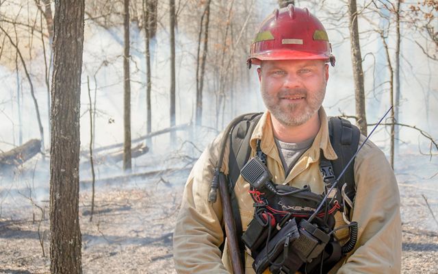 Ryan Gauger smiling for the camera with smoke from a prescribed fire behind him.