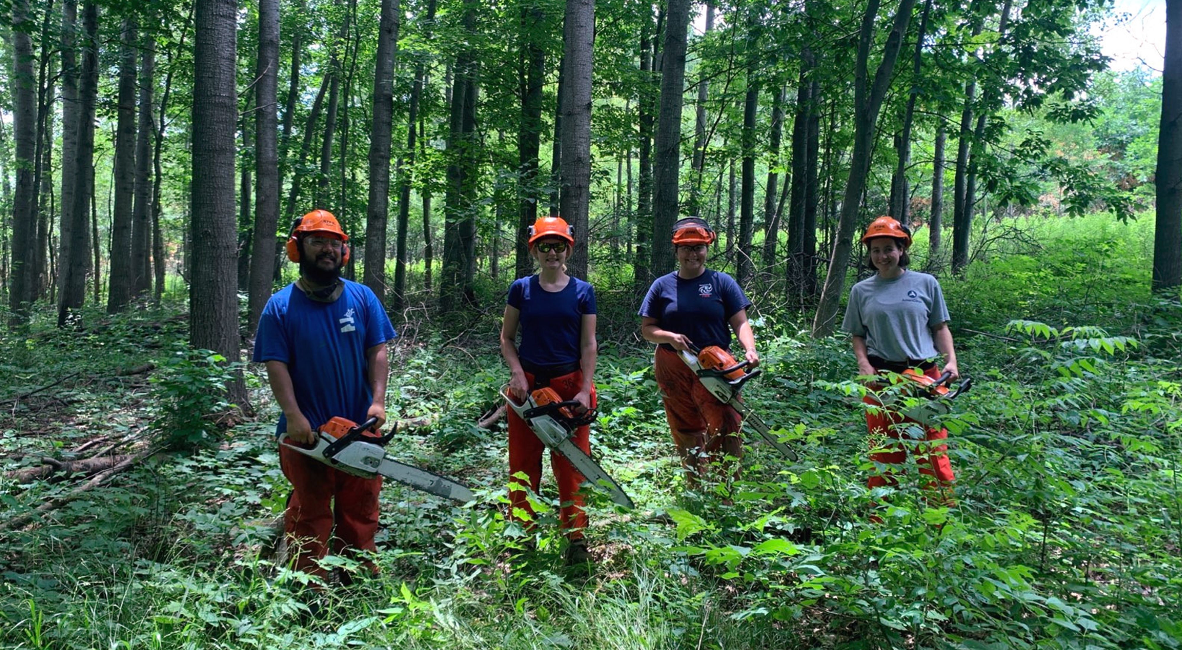 Fie Thao, Gabbi Genz, Karina Cardella and Bobbi Rooney stand in a row in the middle of a green forest, each holding a chainsaw and wearing protective gear, all smiling. 