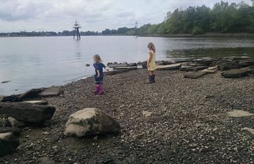 Two young girls wearing rubber boots explore the shoreline of a marsh. One girl has her arm pulled back ready to throw a rock into the water.