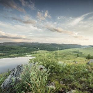 A wide landscape showing rolling green hills, a reflective lake, and dense forest in the distance under a soft, cloudy sky.