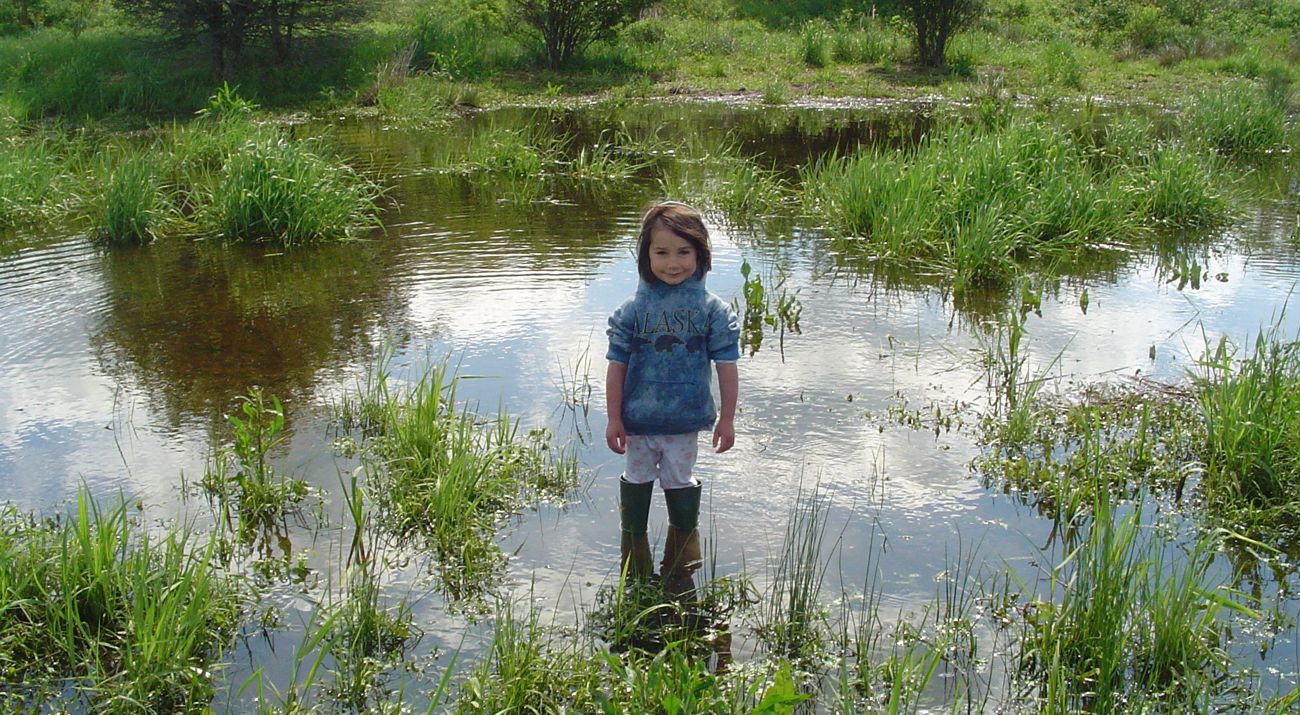 A little girl in rubber boots standing in ankle-deep water with marsh grasses scattered nearby.