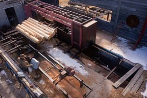 Overhead view of Paul Rust standing at a computer attached to sawmill machinery that has large logs on it.
