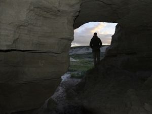 A person stands in a rocky outcrop and looks out onto a prairie through an opening in the crag.