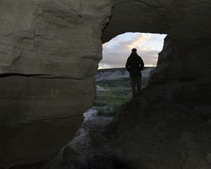 A person stands in a rocky outcrop and looks out onto a prairie through an opening in the crag.