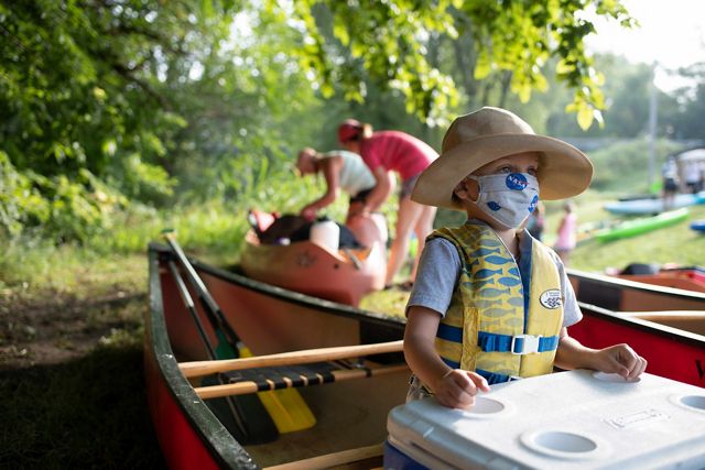 A young boy stands in a docked canoe in front of a cooler. he has on a floppy hat and is wearing a face mask with the logo of NASA on the front. he is wearing a yellow life vest with blue fish design.