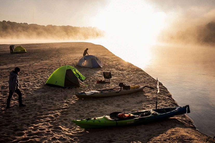 Silhouettes of three campers in fog and early morning light as they move around the campsite along a sandy bank of an island in the river. 