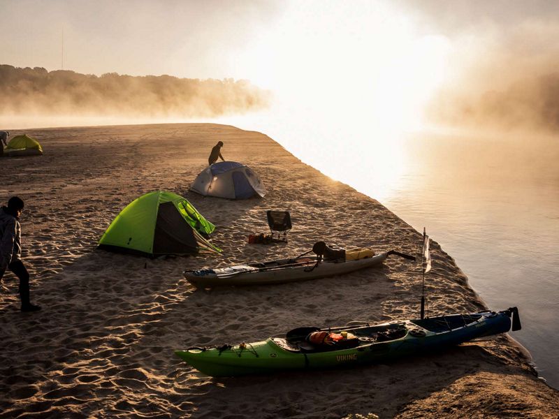 Silhouettes of three campers in fog and early morning light as they move around the campsite along a sandy bank of an island in the river.