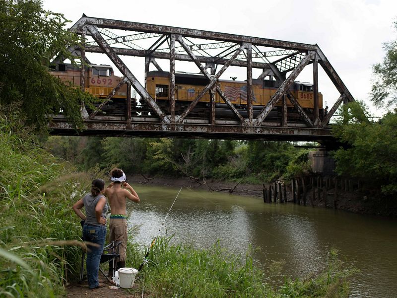 Two individuals stand in grass in front of a body of water and watch as a train passes across a bridge across the water behind them.