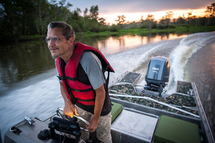 A man stands in a motor boat and holds the steering wheel with a blurry swamp behind him, indicating the speed he is traveling.