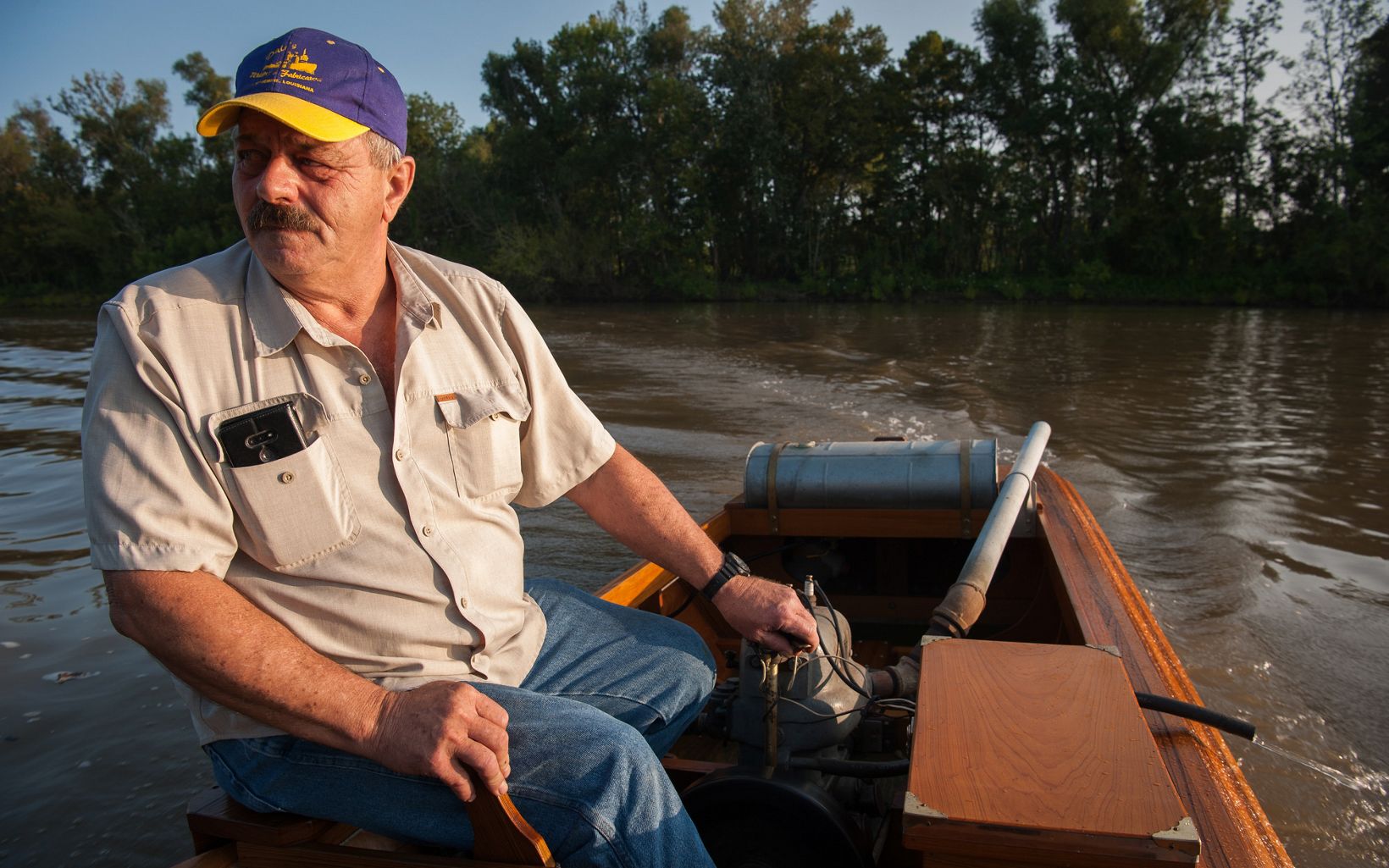 A man in a short-sleeved collared shirt and a baseball hat navigates his wooden boat through swampy water. 
