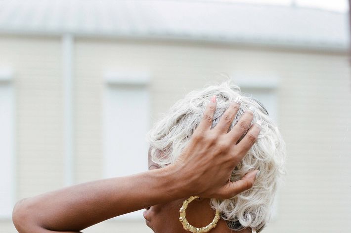 A woman is in the frame from torso up. she is wearing a black tank top and gold hoop earrings. She is in profile but her arm is raised and her hand is touching the back of her head, obscuring her face