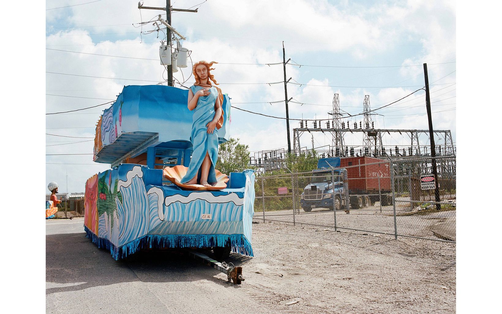 A blue parade float is shot from behind in an industrial space. a statue of Venus is attached at the back of the float and the bottom of the float has details of waves. 