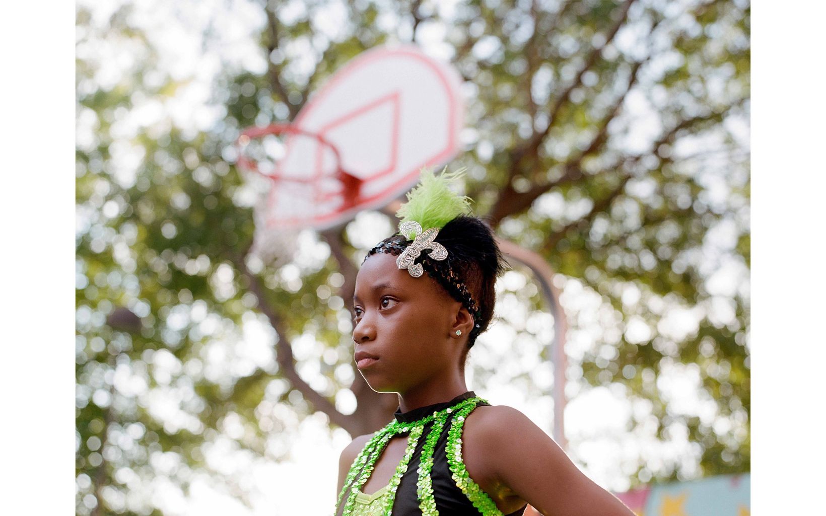 A young girl poses for a portrait. she is wearing a green and black dance leotard and an embellished headband and she stares into the distance left of the frame. A baseketball net is in background.