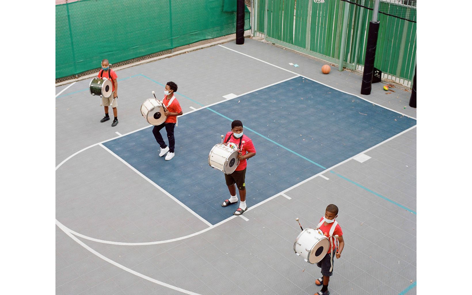 Four young boys in red shirts stand in a line on an outdoor basketball court. They each have a drum strapped to the front of them and are practicing drumming.