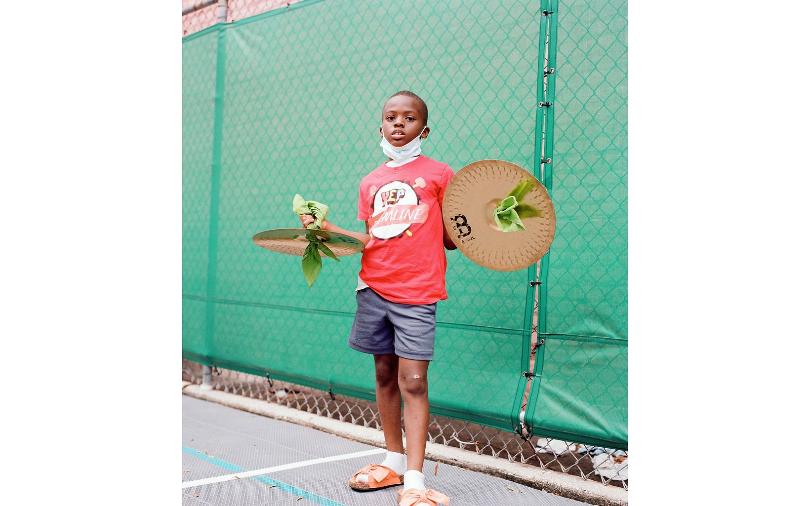 A young boy in a red t-shirt and shorts poses with two cymbals held out to his sides. he is standing outdoors in front of fencing covered in green mesh.
