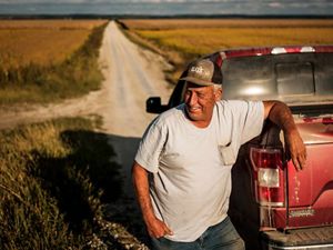 A man wears a baseball hat and t-shirt and stands to the left of his  red truck, his right elbow propped on the top of the cab. A long dirt road and fields of crops are behind him.