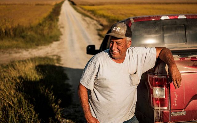 A man wears a baseball hat and t-shirt and stands to the left of his  red truck, his right elbow propped on the top of the cab. A long dirt road and fields of crops are behind him.
