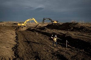 A Man in yellow safety gear and hard hat walks away from heavy machinery in a large plot of land that has been leveled by the machinery.