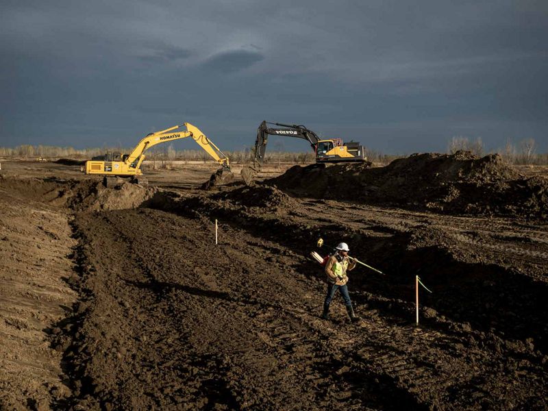 A Man in yellow safety gear and hard hat walks away from heavy machinery in a large plot of land that has been leveled by the machinery.