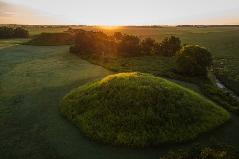 Early morning light illuminates a grassy mound of land surrounded by flat, grassy land. A few trees are surrounding this mount of earth. In the background is a similar, but flat-topped, mound of earth