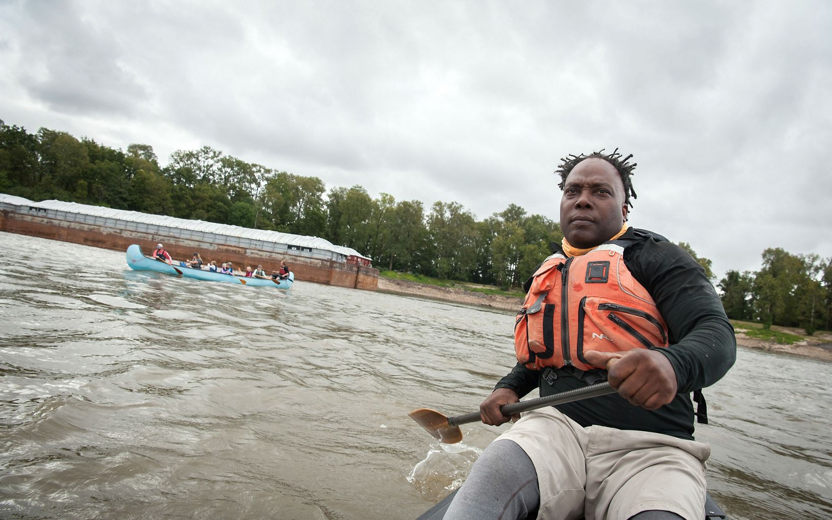 A man paddles his boat in murky water. He is wearing a safety vest and behind him a canoe filled with people and a docked barge are visible. 