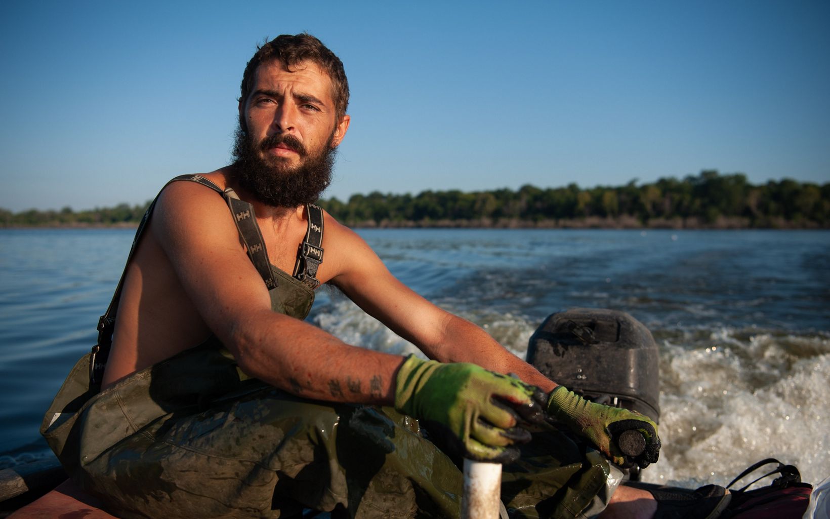A bearded man stares directly at the camera while he steers a motor boat on a body of water. He is wearing fishing overalls.