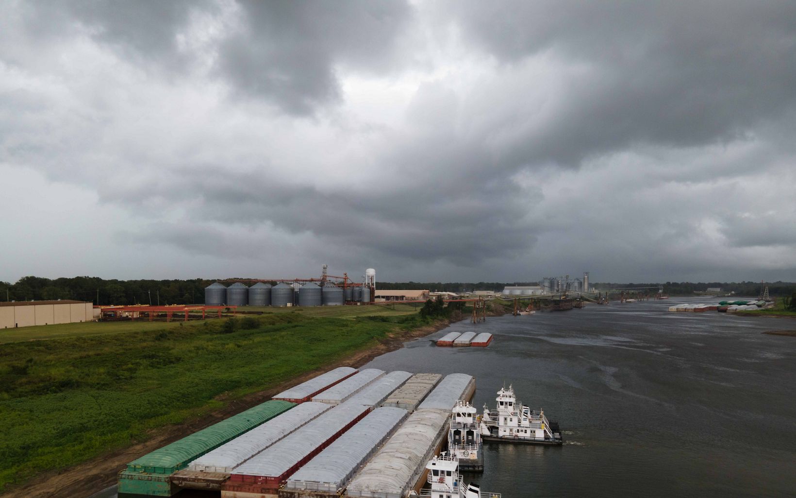 Barges are docked against a river under dark, cloudy skies. in the background are rows of silos and other industrial buildings. 