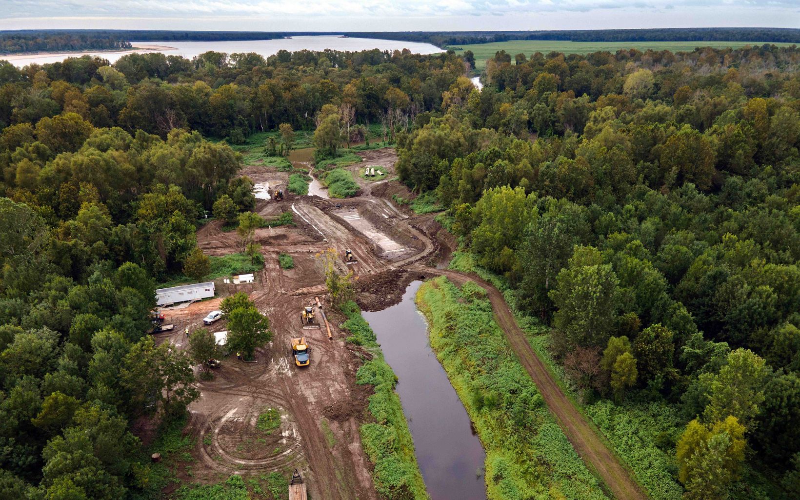 An aerial view of heavy machinery working along a canal of water and surrounded by trees. In the distance a river is visible. The heavy machinery has created swirled patterns tire marks in mud.