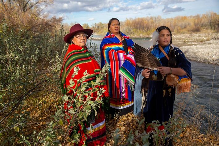 Three generations of women stand within berry bushes at the edge of a creek. They are wearing brightly striped blankets and traditional Blackfeet tribal clothing.