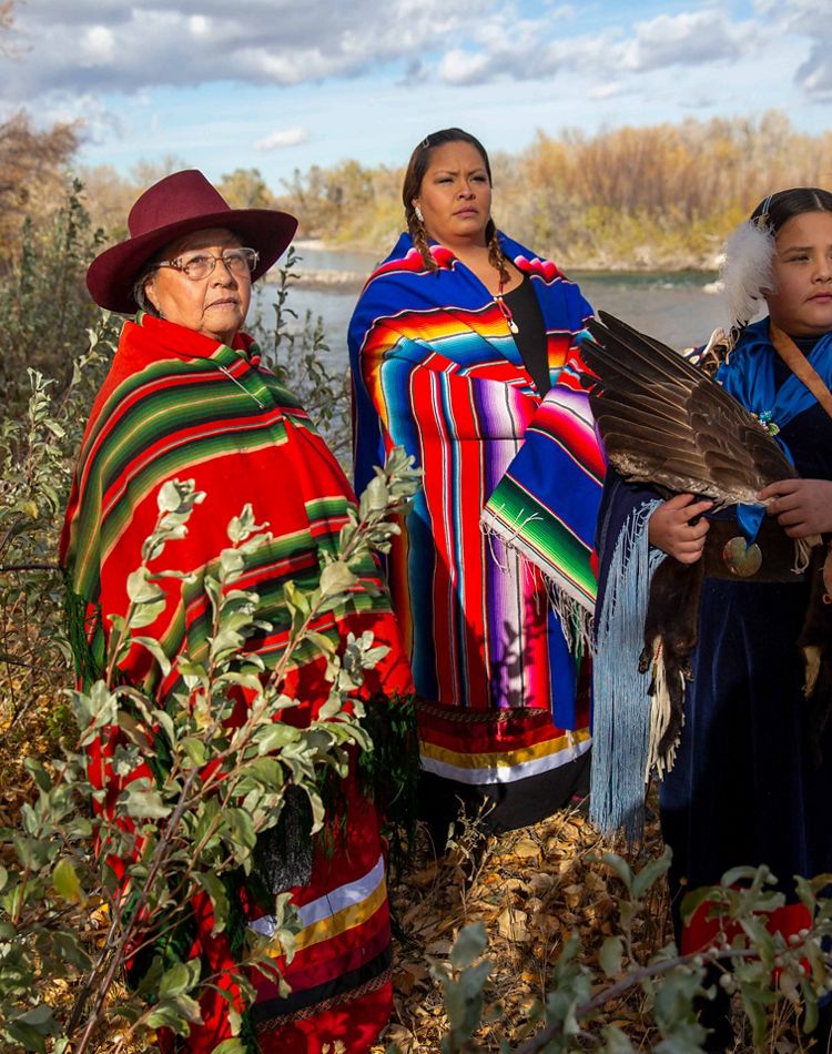 3 women wear richly colored blankets on a river bank.