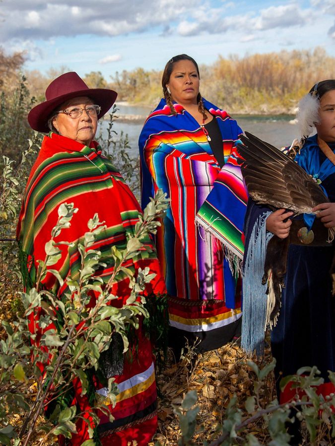 3 women wear richly colored blankets on a river bank.