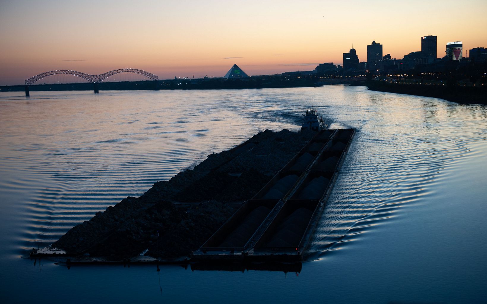 The silhouette of a barge moving down the river, rippling the water around it at sunset. The skyline of the city of Memphis is visible in the distance.