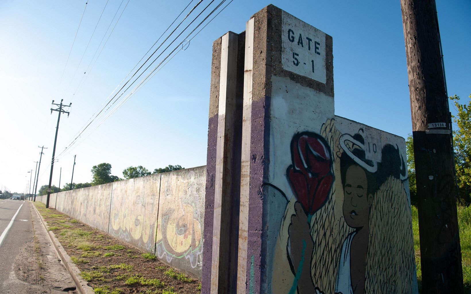 A long wall runs on the right side of a road and is decorated with paintings. At the end of the wall, the painting reads "Gate 5-1" and a depicts an angel with a halo holding a rose. 