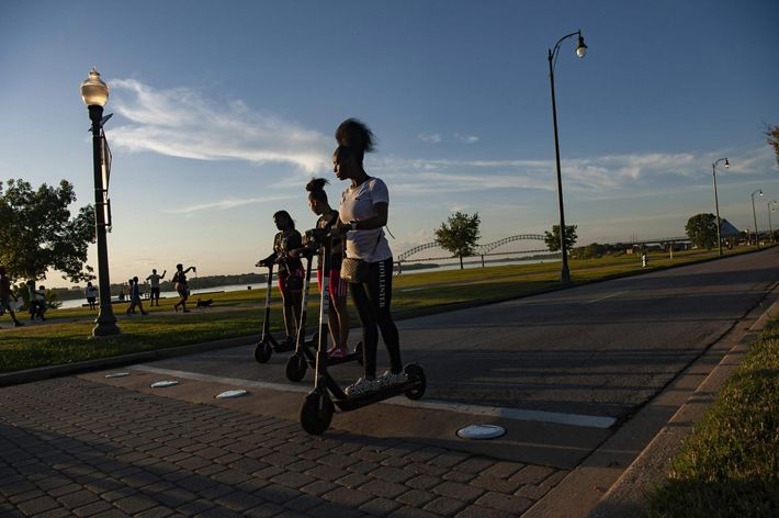 Three teenage girls ride their electric scooters on a paved pedestrian path as the sun sets. Their faces are partly in shadow and light from the angle of the sun.