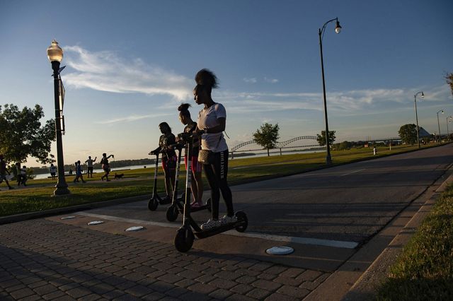 Three teenage girls ride their electric scooters on a paved pedestrian path as the sun sets. Their faces are partly in shadow and light from the angle of the sun. 