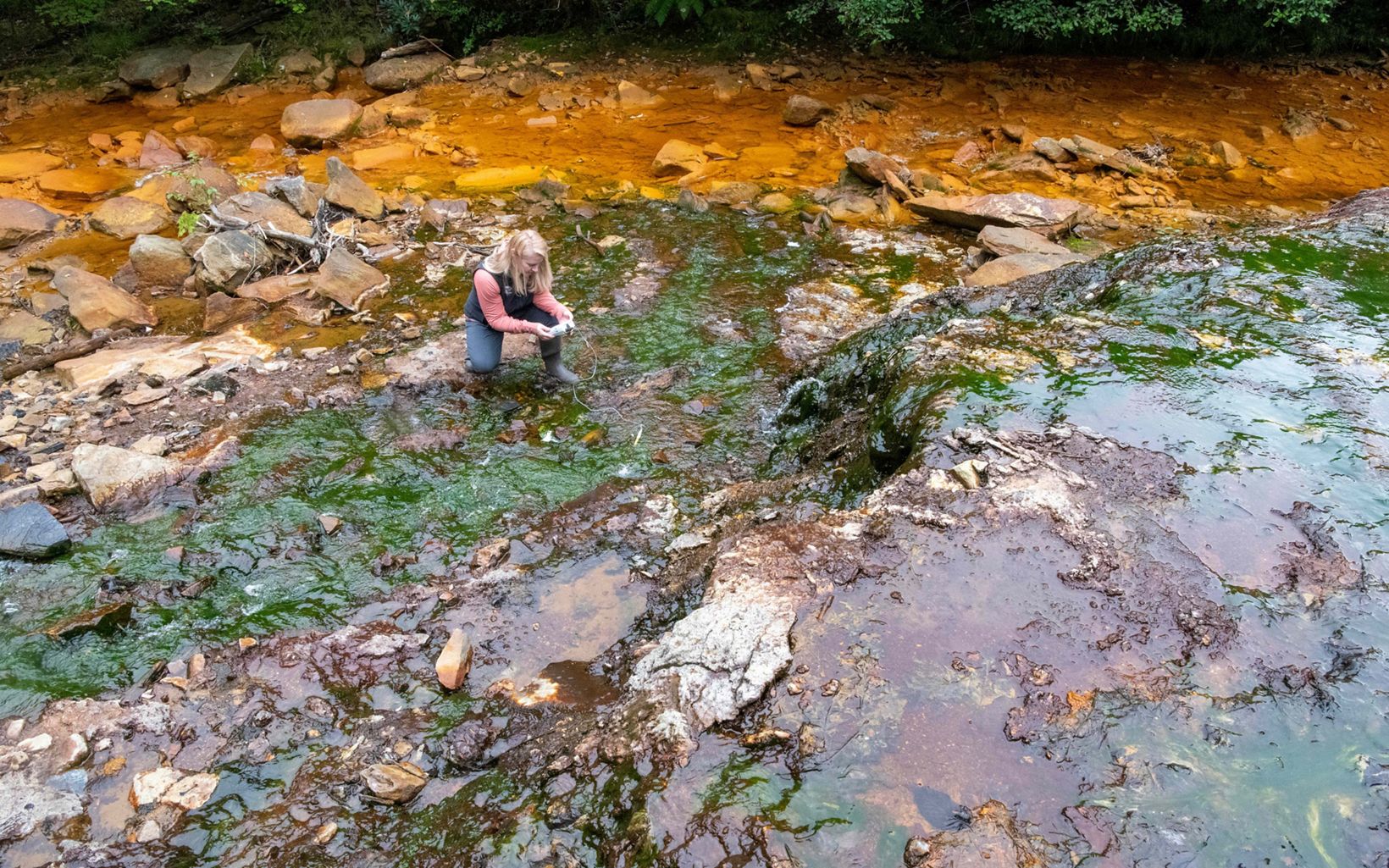 A woman crouches in a shallow part of water that is unnaturally colored green and orange and brown.She has an instrument in her hands.
