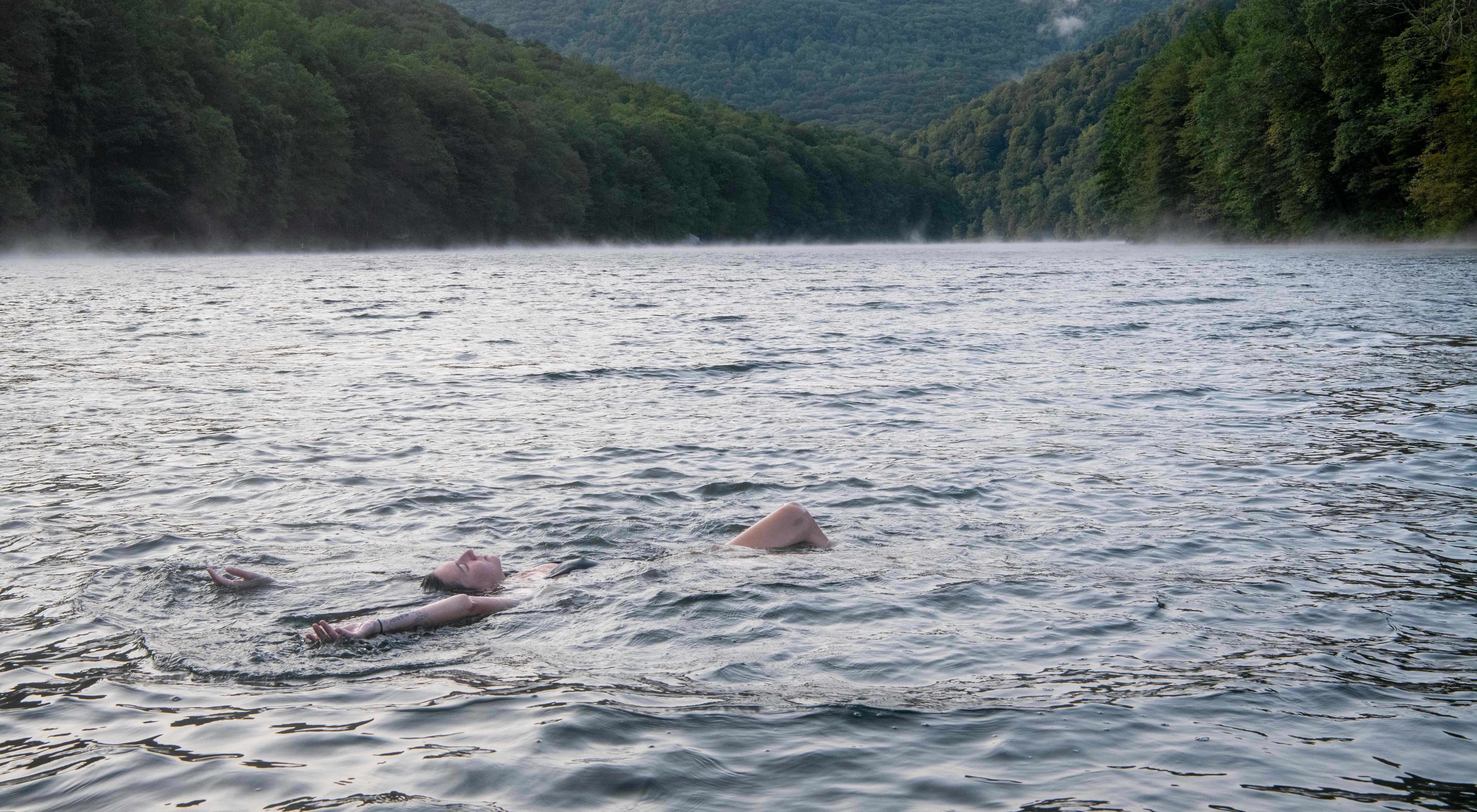 A woman floats face up  in the middle of a large body of water with her arms outstretched in the water. A line of trees is in the background. 