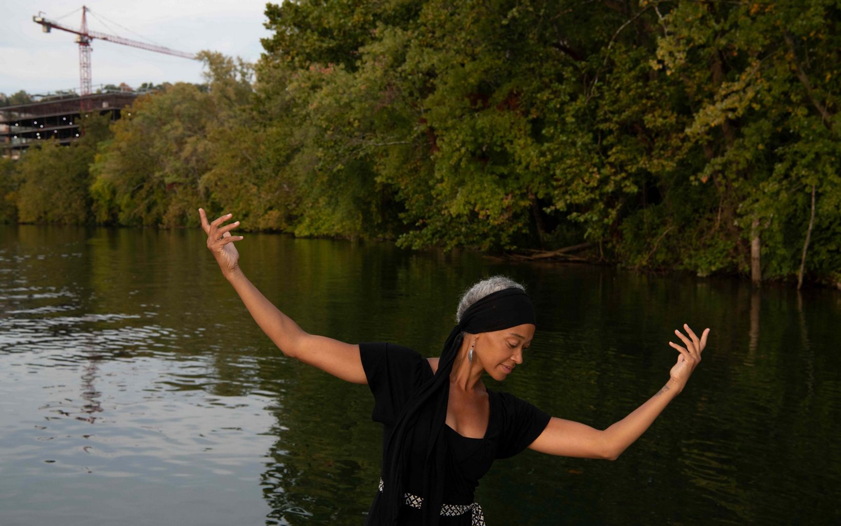 A woman arches her arms into the air in a dance pose with a river and trees in the background. 
