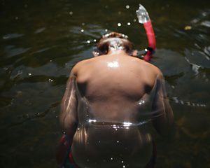 A young boy's back is visible as he uses a snorkel to dip his head into murky water. Sunlight is creating sparkly spots in the water and on his back.