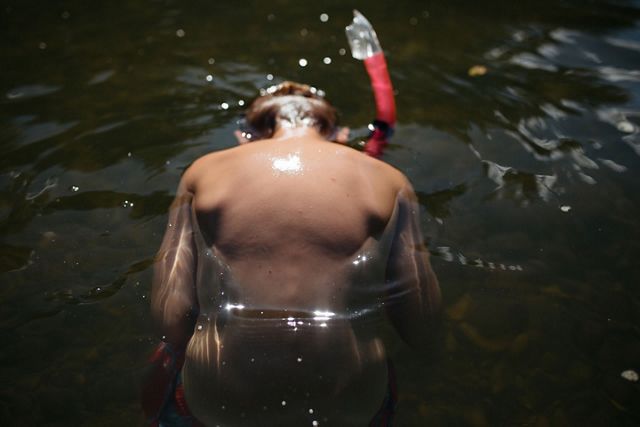 A young boy swims in dark water. His back is visible and a bit of his arms and his neck is visible as he submerges his face into the water. The top of a red snorkel is visible.