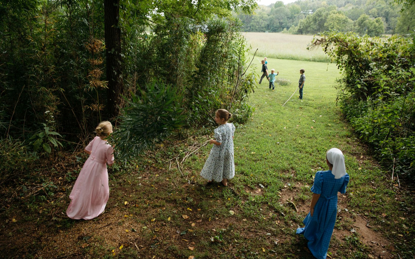 Three girls in pink, floral, and blue handmaid dresses walk away from the camera on a grassy path towards a group of young boys playing with sticks. 