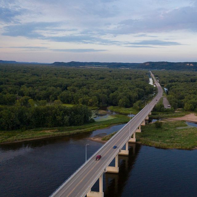 Aerial view of a raised road crossing the Mississippi River near Wabasha, MN. The road leads off into the distance, through dense forested land toward hills in the far distance.