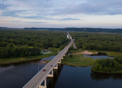 Aerial view of a raised road crossing the Mississippi River near Wabasha, MN. The road leads off into the distance, through dense forested land toward hills in the far distance.