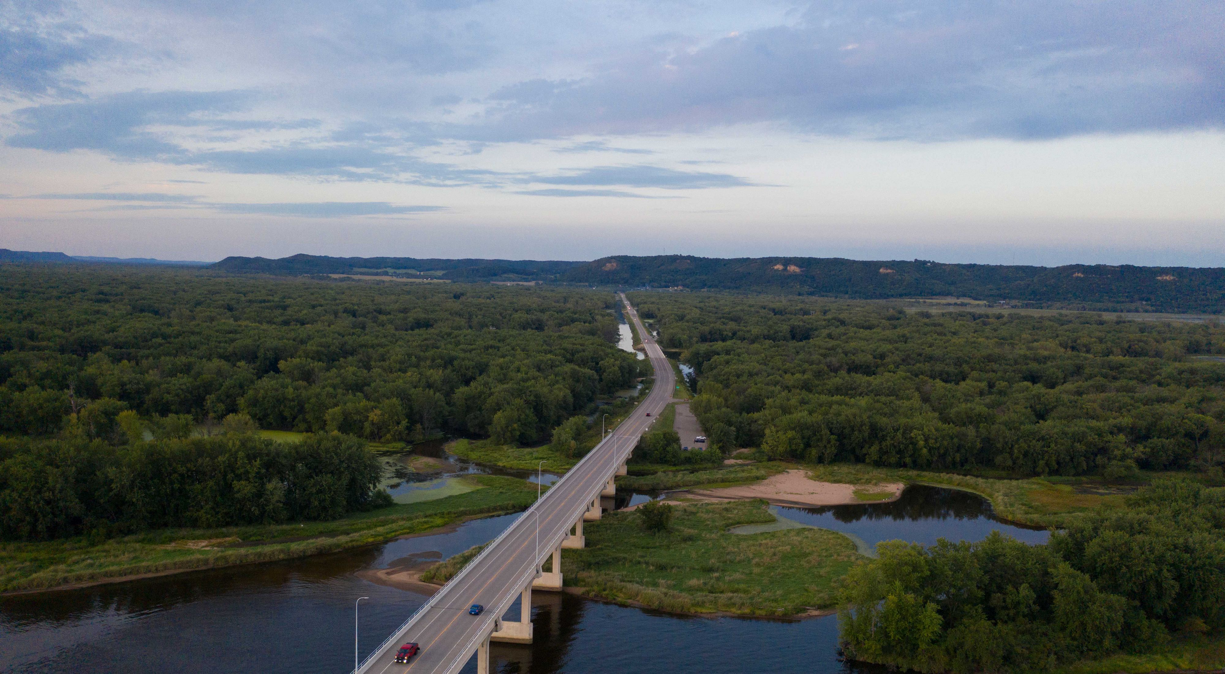 Aerial view of a bridge over the Mississippi River near Wabasha, Minnesota, and a road that extends through thick green forests toward green hills in the distance.