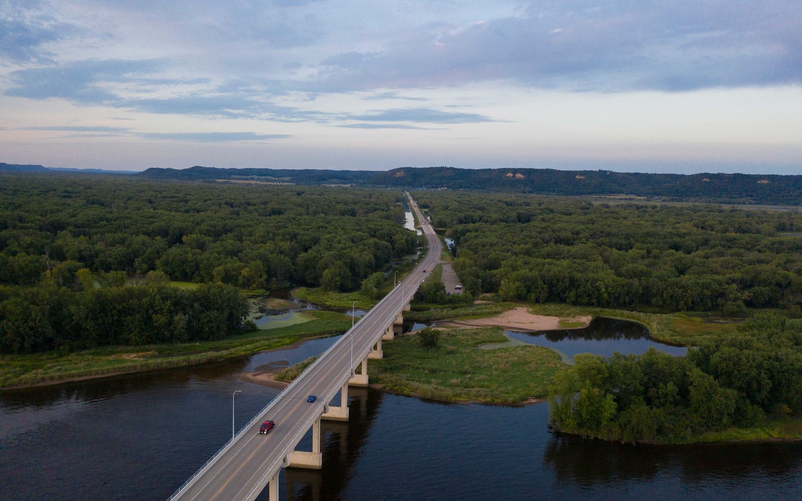 A bridge seen from above with cars driving along it. Below the bridge is blue water and in the foreground is an abundant and green forested area. 