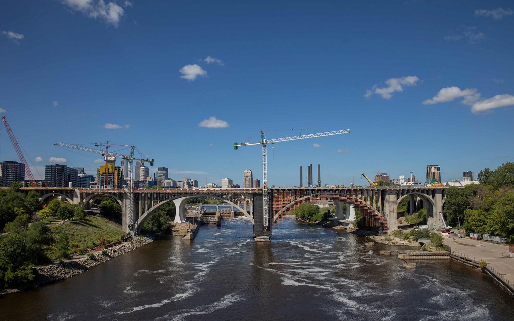 Water flows under a bridge with  multiple cranes in the skyline that are near the bridge. A sunny sky and the skyline of the city are in the background. 
